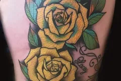 Un tatuaje vibrante neo-tradicional en el muslo con tres grandes rosas amarillas, con hojas verdes marcadas y sombreado detallado. Lo acompaña un tatuaje pequeño de tipo caligrafía con el nombre “Lacey”, con líneas decorativas y remolinos. Tatuaje color por Agu en Hipno Tattoo Estepona.
