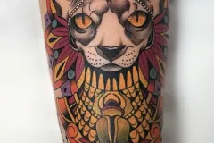 Un tatuaje vibrante neo-tradicional en el brazo que presenta un retrato detallado de un felino con ojos amarillos llamativos. El diseño incorpora motivos egipcios, incluyendo un tocado ornamentado con una cobra y una pieza de escarabajo fúnebre (escarabajo) con incrustaciones de “joyas” y colores vivos en el pecho. Se completa con cadenas negras con cuentas que cuelgan. Tatuaje color por Agu en Hipno Tattoo Estepona.