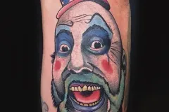 Un tatuaje vibrante y realista en estilo retrato de un hombre calvo con maquillaje de payaso exagerado y un sombrero patriótico rojo, blanco y azul tipo Uncle Sam. El diseño detallado muestra ojos grandes con sombra de ojos azul, mejillas rosadas, la boca abierta y una pajarita roja en la parte inferior.