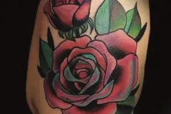 Un primer plano de un tatuaje neo-tradicional en la parte alta del brazo que presenta una gran rosa roja y vibrante, con un capullo de rosa más pequeño y hojas verdes marcadas. El diseño se destaca con contornos negros gruesos y un sombreado en tonos teal (verde azulado) profundo. Tatuaje color por Agu en Hipno Tattoo Estepona.