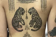 Una fotografía detallada de un tatuaje tradicional tailandés Sak Yant en la espalda, realizado con tinta negra. El diseño presenta una composición grande y simétrica: a la izquierda aparece un tigre a rayas y a la derecha un leopardo con manchas, enfrentados entre sí. Por encima de los animales hay un intrincado Yantra geométrico, junto con columnas verticales de escritura antigua de los Khmer. En la parte baja de la espalda, una flor de loto se sitúa en el centro y está enmarcada por una línea curva de texto de oraciones sagradas. Tatuaje blackwork por Agu en Hipno Tattoo Estepona.