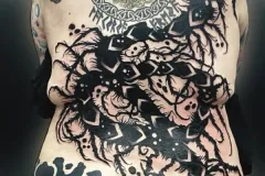 Una vista posterior de la espalda de una persona con un gran conjunto ecléctico de tatuajes en varios estilos. En la parte superior, al centro, hay un tatuaje circular del Árbol de la Vida con un colorido sutil, enmarcado por un intrincado trabajo de nudos celtas. En la zona media e inferior domina una pieza enorme y caótica de blackwork: muestra figuras abstractas, calaveras y patrones geométricos entrelazados con estructuras oscuras que parecen raíces. En la cadera izquierda baja aparece un diseño tribal marcado con grandes remolinos en negro, mientras que en el hombro izquierdo superior se ve un tatuaje de mariposa. Tatuaje blackwork por Agu en Hipno Tattoo Estepona.