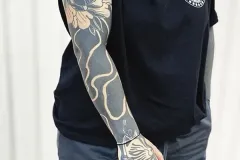 Una vista lateral de una persona que muestra un tatuaje blackwork de manga completa y de gran detalle. En el hombro destaca una gran flor Daruma, con patrones de olas en espacio negativo que bajan por el antebrazo. También se aprecia un tatuaje intrincado de la mano Kitsune. Lleva una camiseta negra de Hipno Tattoo Estepona | Blackwork Club. Tatuaje blackwork por Agu en Hipno Tattoo Estepona.