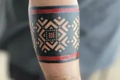 Un primer plano del antebrazo que muestra un tatuaje tipo brazalete geométrico y marcado. El diseño presenta una banda negra gruesa con un patrón simétrico y repetitivo de rombos y zigzag en el espacio negativo, enmarcado por líneas finas de color rojo oscuro en la parte superior e inferior. Tatuaje blackwork por Agu en Hipno Tattoo Estepona.