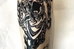 Un primer plano de la pierna baja muestra un tatuaje grande y marcado en bold blackwork en la pantorrilla. El diseño de alto contraste presenta arriba una máscara de demonio feroz y con cuernos (tipo oni), que se transforma en una boca abierta y surreal, y desciende hacia abajo convirtiéndose en garras irregulares con aspecto de llamas, junto con formas orgánicas abstractas. Tatuaje blackwork por Agu en Hipno Tattoo Estepona.