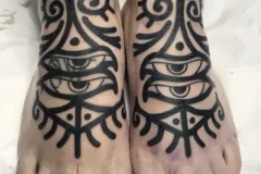 Una vista desde arriba de un par de pies con tatuajes a juego en bold blackwork. El diseño cubre la parte superior de los pies y se extiende hacia arriba por los tobillos. En el centro hay un motivo realista de un ojo, rodeado de patrones abstractos tribales, con remolinos, puntos y líneas radiales. En el tobillo derecho se aprecia además una banda negra sólida. Tatuaje blackwork por Agu en Hipno Tattoo Estepona.