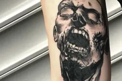 Un primer plano de la parte superior del brazo con un tatuaje hiperrealista en negro y gris, muy detallado, que representa una cara de zombi gritando. El diseño de terror muestra un rostro cadavérico y esquelético, con la boca muy abierta y un sombreado oscuro y marcado. Tatuaje blackwork por Agu en Hipno Tattoo Estepona.