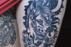 Un tatuaje detallado en blackwork en el muslo con la cabeza de un unicornio, adornada con un intrincado arnés o bozal tipo brida con apariencia de joyas. El diseño está enmarcado en el fondo por una luna creciente y, en la parte inferior, por rosas en plena floración. Todo está realizado en tinta negra, con un estilo oscuro e ilustrativo. Tatuaje blackwork por Agu en Hipno Tattoo Estepona.