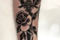 Un primer plano del antebrazo muestra un tatuaje neo-tradicional detallado en negro y gris con una daga vertical. Una gran rosa en plena floración, con hojas y tallos intrincados, envuelve el mango y la hoja, con contornos marcados y sombreado suave. Tatuaje blackwork por Agu en Hipno Tattoo Estepona.