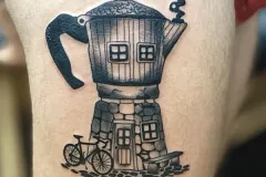 A detailed black and grey illustrative tattoo on a thigh depicting a whimsical Moka pot espresso maker transformed into a cozy house. The design features a stone foundation with a door and window, upper-level windows, a bicycle parked outside, and smoke billowing from the spout. Tatuaje blackwork por Agu en Hipno Tattoo Estepona.