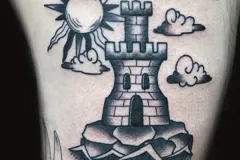 Un tatuaje ilustrativo en negro y gris que muestra una torre de castillo de piedra con almenas, colocada sobre una base rocosa. Desde lo alto de la torre sale una bandera que dice “LIBERTAD”, mientras que alrededor del edificio aparecen un sol tradicional y nubes estilizadas, en un estilo gráfico y marcado. Tatuaje blackwork por Agu en Hipno Tattoo Estepona.