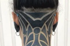 Una vista posterior de una persona con el cabello oscuro recogido en un moño alto, mostrando una nuca rapada con un tatuaje blackwork geométrico y atrevido en el cuello. El diseño simétrico utiliza líneas negras gruesas y espacio negativo para crear patrones abstractos inspirados en el estilo tribal. La persona también tiene los lóbulos de las orejas estirados, con plugs negros y joyería en forma de anillo plateado. Tatuaje blackwork por Agu en Hipno Tattoo Estepona.