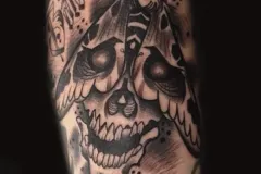 Un tatuaje detallado en negro y gris que muestra un cráneo humano con una gran polilla posada sobre la frente. El diseño incluye un sombreado intrincado, salpicaduras con dotwork dispersas y letras góticas irregulares flotando sobre el insecto. Tatuaje blackwork por Agu en Hipno Tattoo Estepona.
