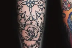 Un tatuaje vertical en negro y gris en una pierna que presenta un mandala floral intrincado con un nudo celta central en la parte superior. El diseño se va prolongando hacia abajo en una rosa detallada con sombreado, y termina con un diseño geométrico que incluye un nudo triquetra en la base. Tatuaje blackwork por Agu en Hipno Tattoo Estepona.