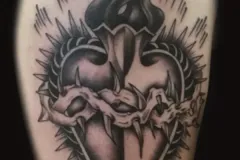 Un tatuaje detallado en negro y gris de un corazón sagrado de estilo estilizado, envuelto en una corona de espinas afiladas. De la parte superior del corazón surgen llamas oscuras y en movimiento; el corazón está enmarcado por un halo espigado con forma de espinas y por líneas radiales que irradian hacia afuera. El diseño incluye un efecto dramático de sombreado dividido en dos tonos. Tatuaje blackwork por Agu en Hipno Tattoo Estepona.