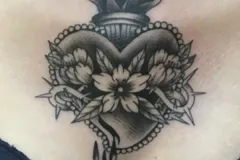 Un tatuaje ilustrativo detallado en negro y gris en el pecho que presenta un diseño de corazón sagrado. El corazón está adornado con flores en plena floración, espinas afiladas y un borde con cuentas. En la parte superior lleva una especie de recipiente en llamas que contiene un corazón pequeño, y se completa con tres gotas que caen desde el punto inferior. Tatuaje blackwork por Agu en Hipno Tattoo Estepona.
