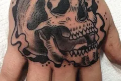 Un tatuaje audaz en negro y gris en la mano con un cráneo grande y detallado, con un efecto de derretido que gotea. Está rodeado por bordes ondulados con apariencia de humo y pequeños acentos de dotwork (puntillismo). Tatuaje blackwork por Agu en Hipno Tattoo Estepona.