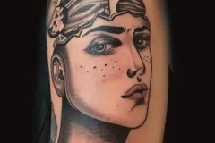 Un tatuaje ilustrativo detallado en negro y gris en el brazo que muestra el retrato del rostro y el cuello de una mujer. La parte superior de su cabeza está abierta con aspecto de concha agrietada, revelando una escena cósmica en el interior, con cuerpos celestes flotando, incluyendo un planeta prominente con anillos. Tatuaje blackwork por Agu en Hipno Tattoo Estepona.