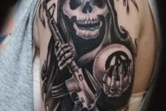 Un tatuaje detallado en negro y gris en el hombro que muestra un cráneo realista de la Muerte (Grim Reaper) sosteniendo un rifle de asalto y un símbolo circular de anarquía con una mano esquelética. Todo está enmarcado por una capa oscura con capucha y una hoja de guadaña. Tatuaje blackwork por Agu en Hipno Tattoo Estepona.