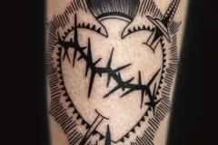 Un primer plano de una pierna con un tatuaje bold de blackwork con un corazón envuelto en alambre de púas o espinas. El diseño está coronado con una llama estilizada, y a ambos lados tiene espadas que lo atraviesan o lo flanquean. Está rodeado por líneas radiales y se completa con pequeñas gotas oscuras tipo lágrimas que caen desde el punto inferior. Tatuaje blackwork por Agu en Hipno Tattoo Estepona.