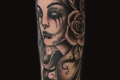 Un tatuaje detallado en negro y gris en el antebrazo que muestra un retrato de medio rostro de una mujer con pequeños cuernos de diablo y una rosa grande. Un lado del rostro es realista, mientras que el otro se transforma en un cráneo con labios cosidos y un ojo oscuro que gotea. Tatuaje blackwork por Agu en Hipno Tattoo Estepona.