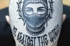 Una vista desde arriba de una cabeza rapada con un gran tatuaje en negro y gris de un rostro que lleva una capucha tipo pasamontañas, enmarcado por una corona de alambre de púas. Debajo del retrato, la frase “ME AGAINST THE WORLD” está tatuada con una tipografía bold gótica blackletter a lo largo de la parte posterior de la cabeza. Tatuaje blackwork por Agu en Hipno Tattoo Estepona.